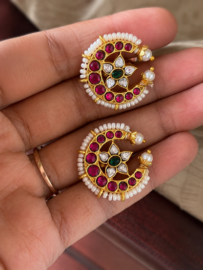 Close-up of a pair of traditional South Indian Chandra Bugudi gold ear cuffs held in a hand, featuring a crescent moon shape lined with tiny white pearls, embedded rubies, and a central star-shaped stone flower motif.