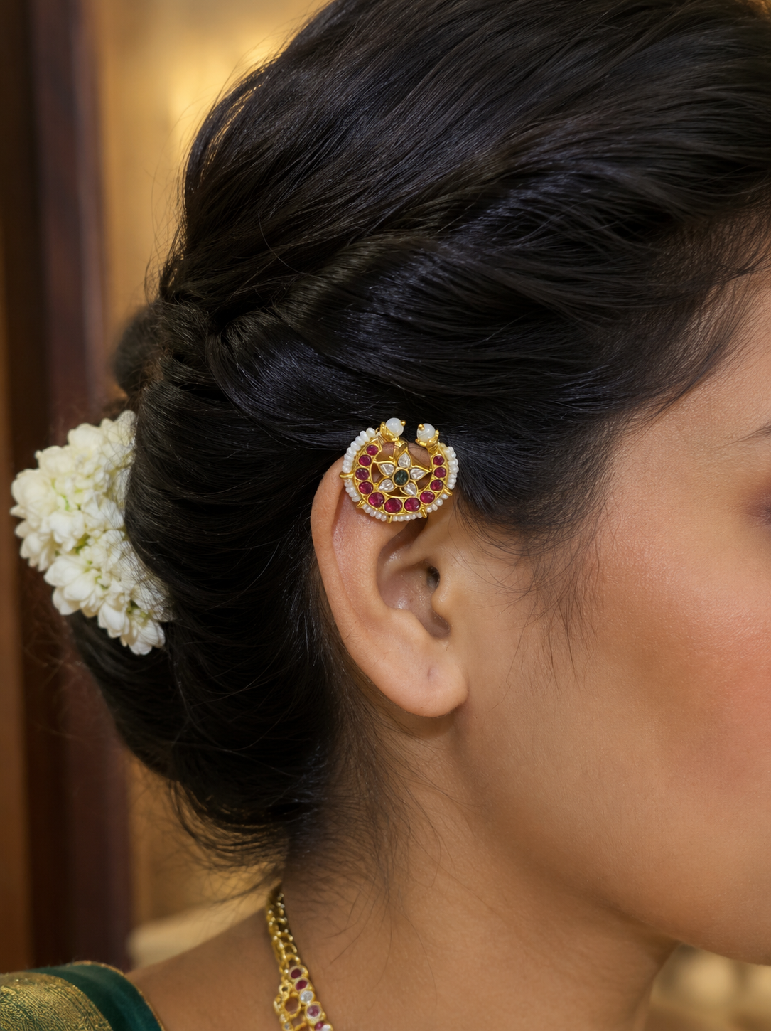 A side profile view of a woman wearing the traditional gold Chandra Bugudi ornament on the upper rim of her ear cartilage, showcasing the ruby, pearl, and kundan star details against a braided hairstyle adorned with jasmine flowers.