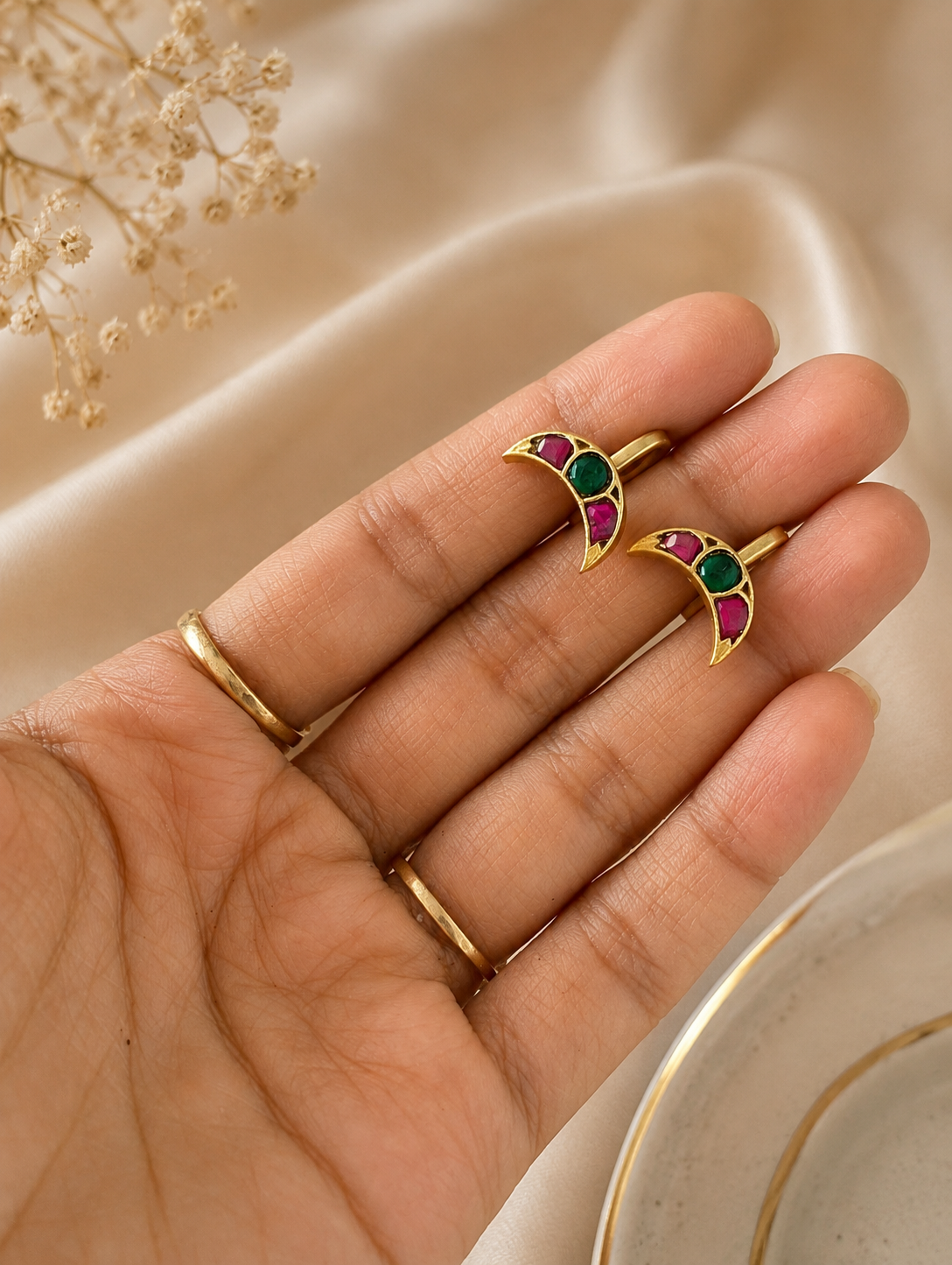 Close-up of a hand holding a pair of gold-toned, crescent moon-shaped Jadau Kundan ear cuffs inlaid with alternating pink rubies and a central green emerald stone.