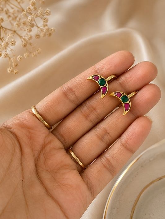 Close-up of a hand holding a pair of gold-toned, crescent moon-shaped Jadau Kundan ear cuffs inlaid with alternating pink rubies and a central green emerald stone.