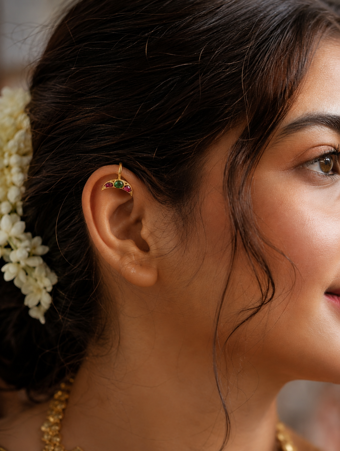 A close-up side profile of a woman wearing a gold crescent moon bugudi ear cuff with ruby and emerald stones on her upper ear cartilage, styled with a soft bridal look and fresh jasmine flowers in her hair.