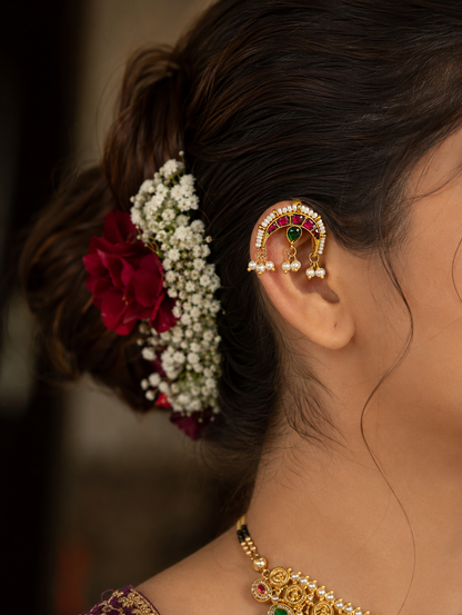 A side profile view of a woman wearing a gold-toned Chandra style bugudi ear cuff on the upper rim of her ear cartilage, beautifully coordinating with a braided updo adorned with fresh white baby's breath and a red rose.
