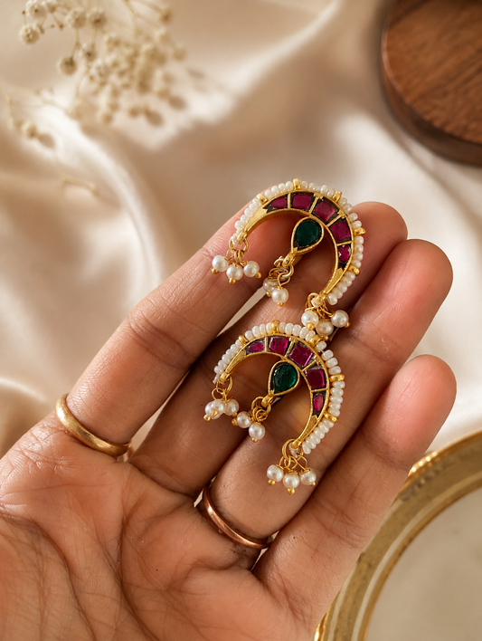 A close-up view of a person's hand holding a pair of traditional gold-toned Chandra style bugudi ear cuffs, showing the crescent silhouette framed by tiny seed pearls, square-cut rubies, a green pear-shaped drop stone, and delicate hanging pearl clusters.