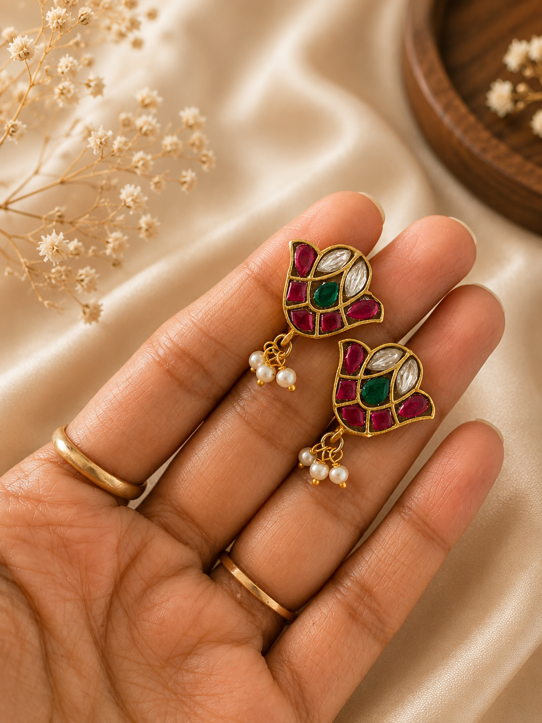 A close-up view of a hand holding a pair of traditional gold-toned Jadau Kundan bugudi ear cuffs designed in a stylized floral bud shape, featuring inlaid red rubies, white kundan stones, a central green emerald, and tiny hanging pearl drops.