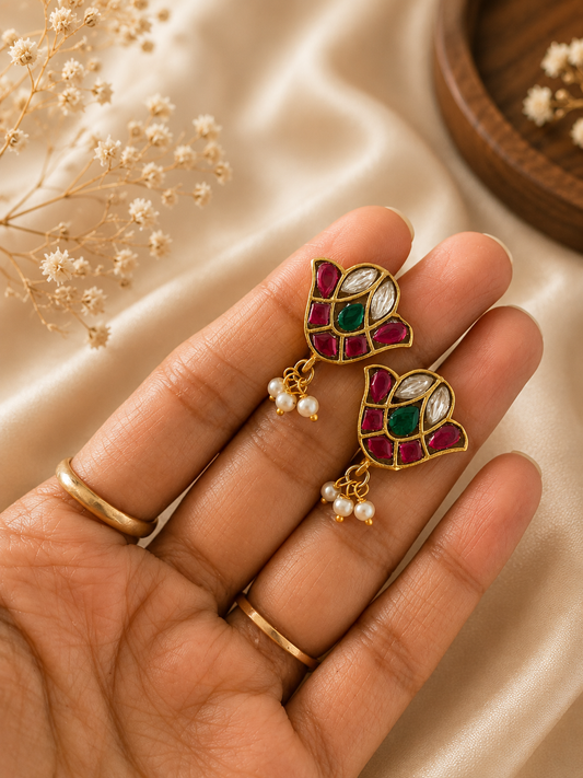A close-up view of a hand holding a pair of traditional gold-toned Jadau Kundan bugudi ear cuffs designed in a stylized floral bud shape, featuring inlaid red rubies, white kundan stones, a central green emerald, and tiny hanging pearl drops.