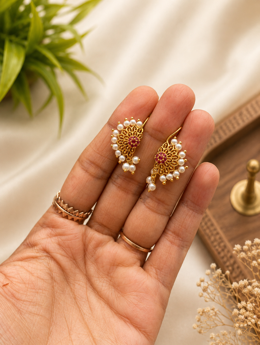 Pair of traditional gold-plated Pearl Style Bugudi earrings with intricate filigree detailing, a central cluster of red ruby stones, and a border of small white pearls, displayed on a hand.