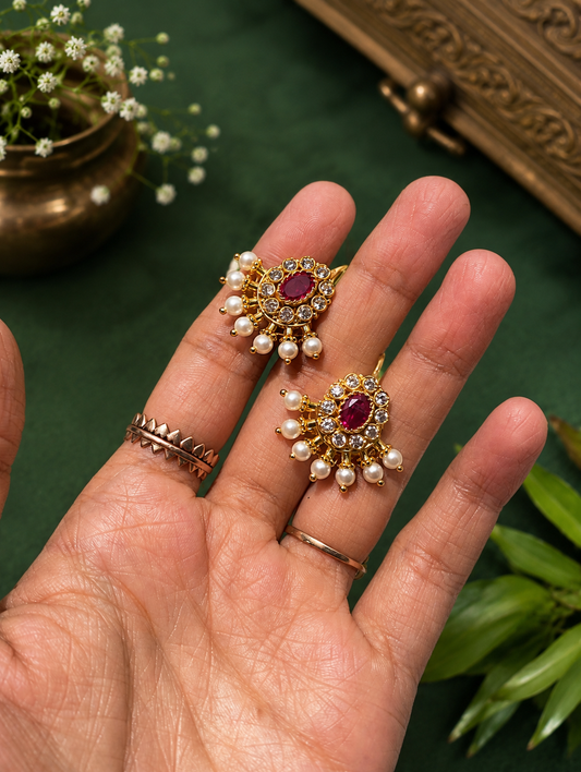 A pair of gold-plated Ruby Pearl Buggudi earrings featuring an oval red ruby stone surrounded by white cubic zirconia and hanging seed pearls, held in a hand.