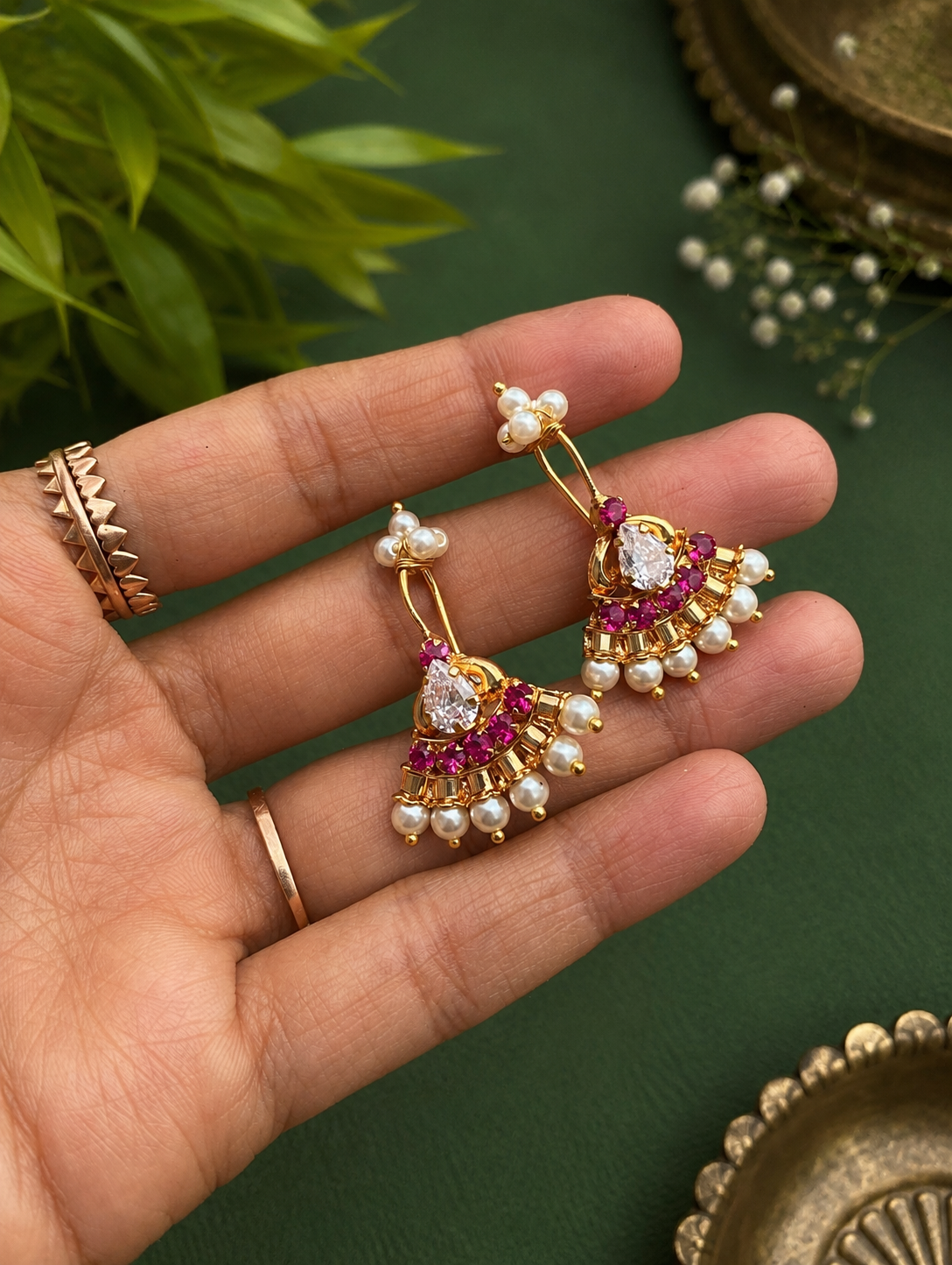 A pair of gold-plated fan-shaped traditional Pearl Ruby Bugudi earrings featuring central pear-shaped CZ stones, a row of ruby-red gems, and a border of small white pearls, held on a hand.