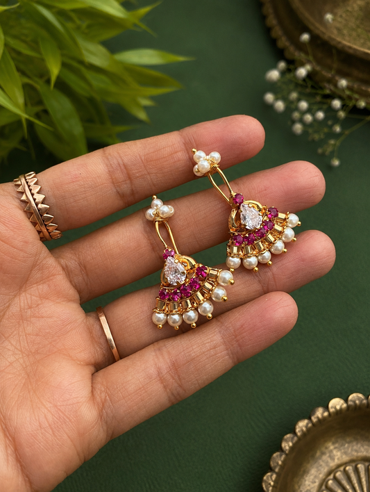 A pair of gold-plated fan-shaped traditional Pearl Ruby Bugudi earrings featuring central pear-shaped CZ stones, a row of ruby-red gems, and a border of small white pearls, held on a hand.