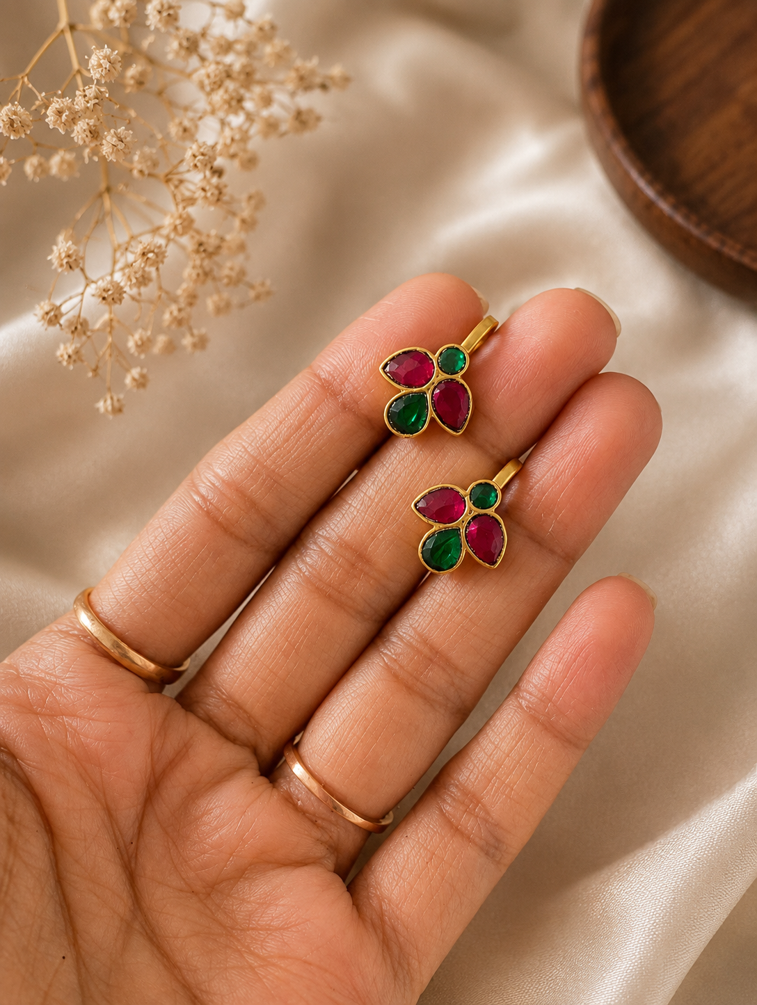 A pair of traditional Leaf Style Jadau Bugudi non-pierced ear cuffs held on a person's hand, showing the intricate gold-plated four-leaf pattern with pink and green stones against a soft satin background.