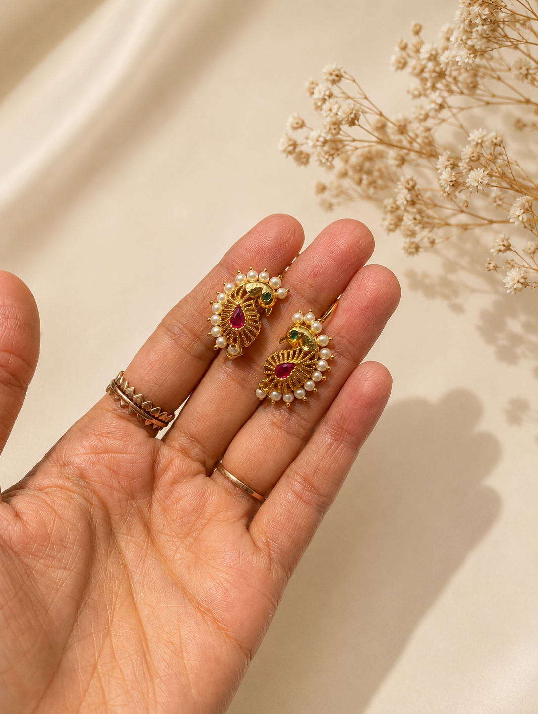 A close-up shot of a pair of ethnic gold-toned peacock-shaped ear cuffs held in the palm of a hand, featuring intricate textured metalwork, a teardrop ruby center, a small green stone eye, and a border of small white pearls.