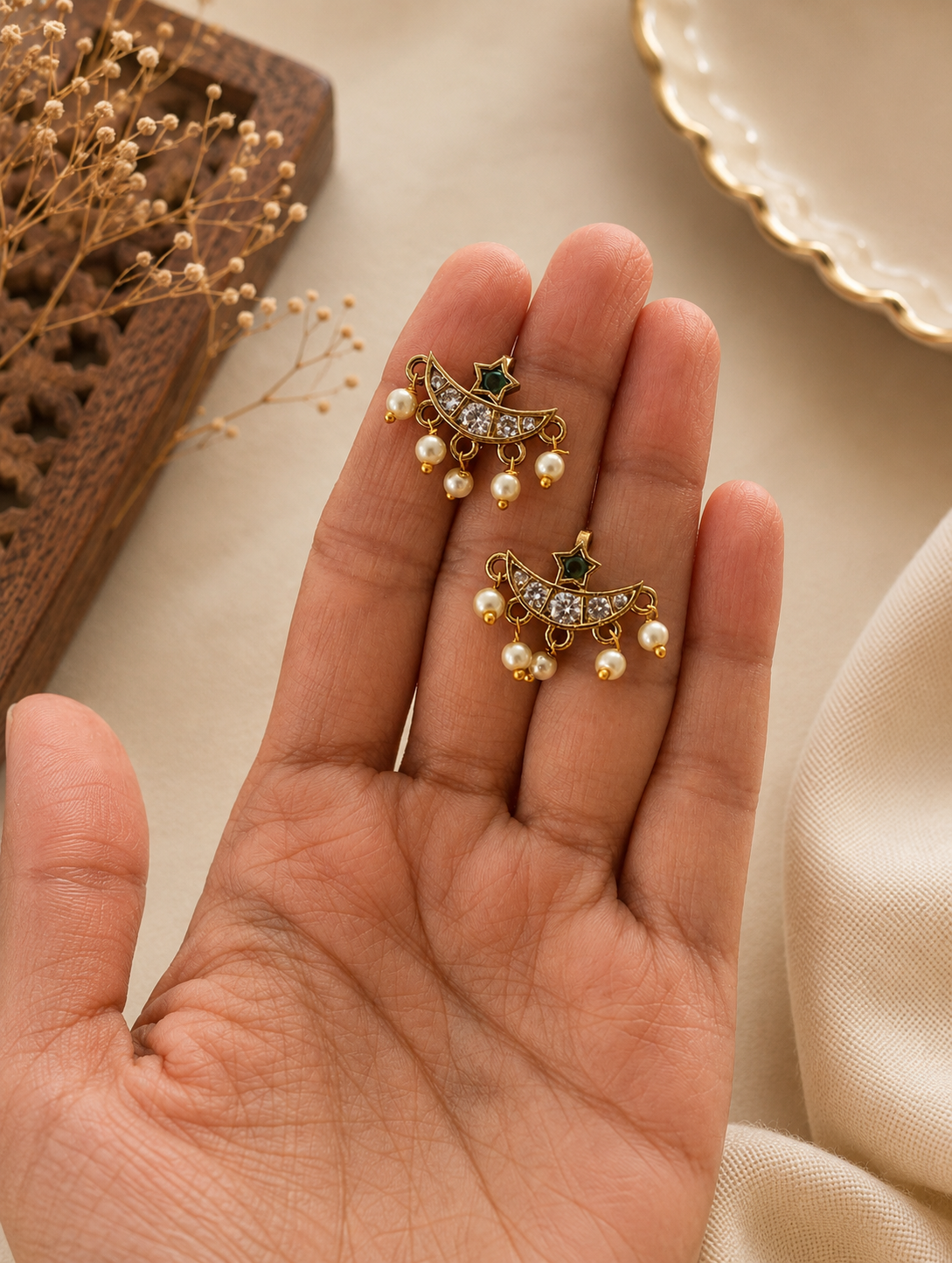 A pair of traditional gold-toned crescent-shaped Bugudi ear cuffs held in a hand, featuring a curved band lined with clear crystals, a tiny green star accent at the top, and five hanging seed pearl drops.