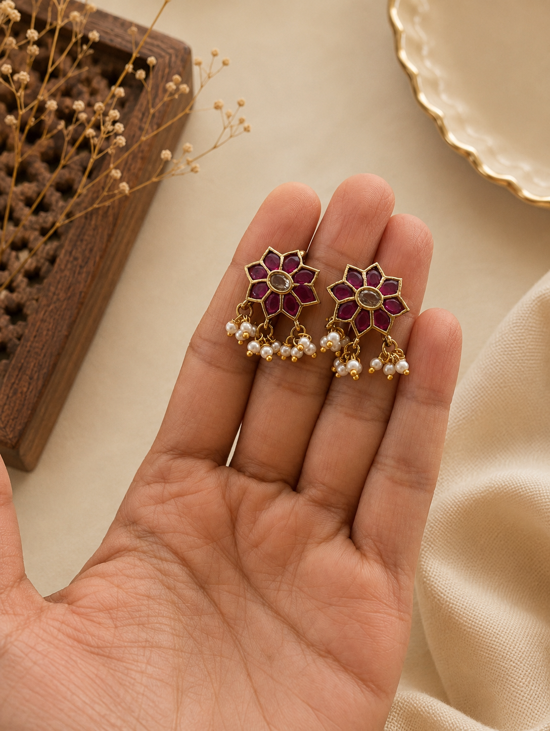 A close-up view of a pair of traditional floral-shaped gold-toned Bugudi ear cuffs held in the palm of a hand, featuring an eight-petal star design inlaid with pink-ruby stones, a clear center crystal, and a dangling fringe of tiny white seed pearls.