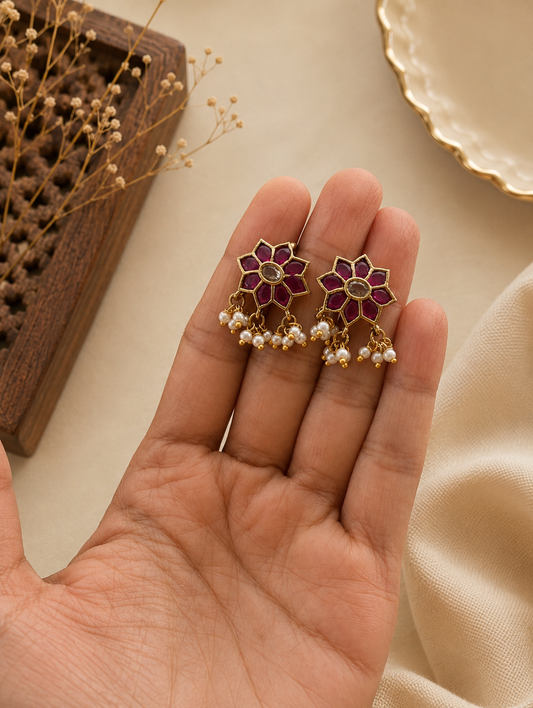 A close-up view of a pair of traditional floral-shaped gold-toned Bugudi ear cuffs held in the palm of a hand, featuring an eight-petal star design inlaid with pink-ruby stones, a clear center crystal, and a dangling fringe of tiny white seed pearls.
