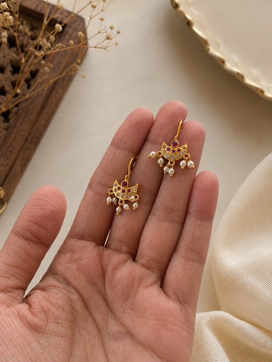 A close-up shot of a pair of traditional gold-toned Bugudi helix earrings held in a hand, featuring a crescent-shaped base encrusted with small clear stones, a central ruby-pink stone, and dangling tiny seed pearls, displayed against a warm, neutral fabric background.