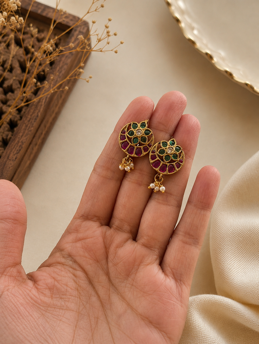 A pair of traditional gold-toned Jadau Bugudi ear cuffs held in a hand, featuring a circular floral motif inlaid with green stones, a clear center crystal, a ruby-pink stone crescent border, and dangling tiny seed pearls.