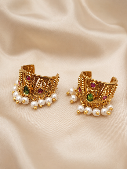 A pair of traditional Indian gold-plated Bugudi earrings for the upper ear cartilage, featuring intricate gold filigree, embedded ruby-red and teardrop green kemp stones, and a fringe of delicate white pearls at the bottom, displayed on a soft, cream-colored satin fabric.
