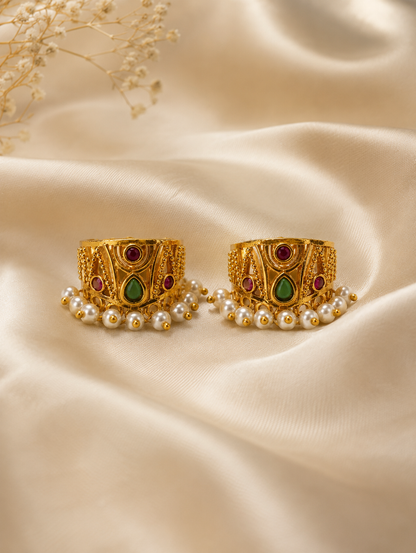 A pair of traditional Indian gold-plated Bugudi earrings for the upper ear cartilage, featuring intricate gold filigree, embedded ruby-red and teardrop green kemp stones, and a fringe of delicate white pearls at the bottom, displayed on a soft, cream-colored satin fabric.