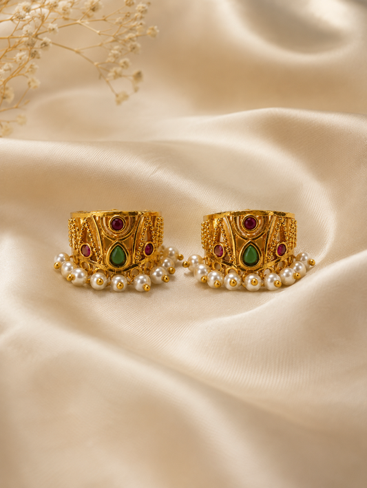 A pair of traditional Indian gold-plated Bugudi earrings for the upper ear cartilage, featuring intricate gold filigree, embedded ruby-red and teardrop green kemp stones, and a fringe of delicate white pearls at the bottom, displayed on a soft, cream-colored satin fabric.