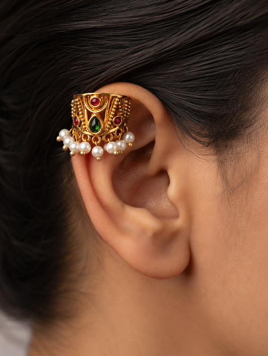 A pair of traditional Indian gold-plated Bugudi earrings for the upper ear cartilage, featuring intricate gold filigree, embedded ruby-red and teardrop green kemp stones, and a fringe of delicate white pearls at the bottom, displayed on a soft, cream-colored satin fabric.