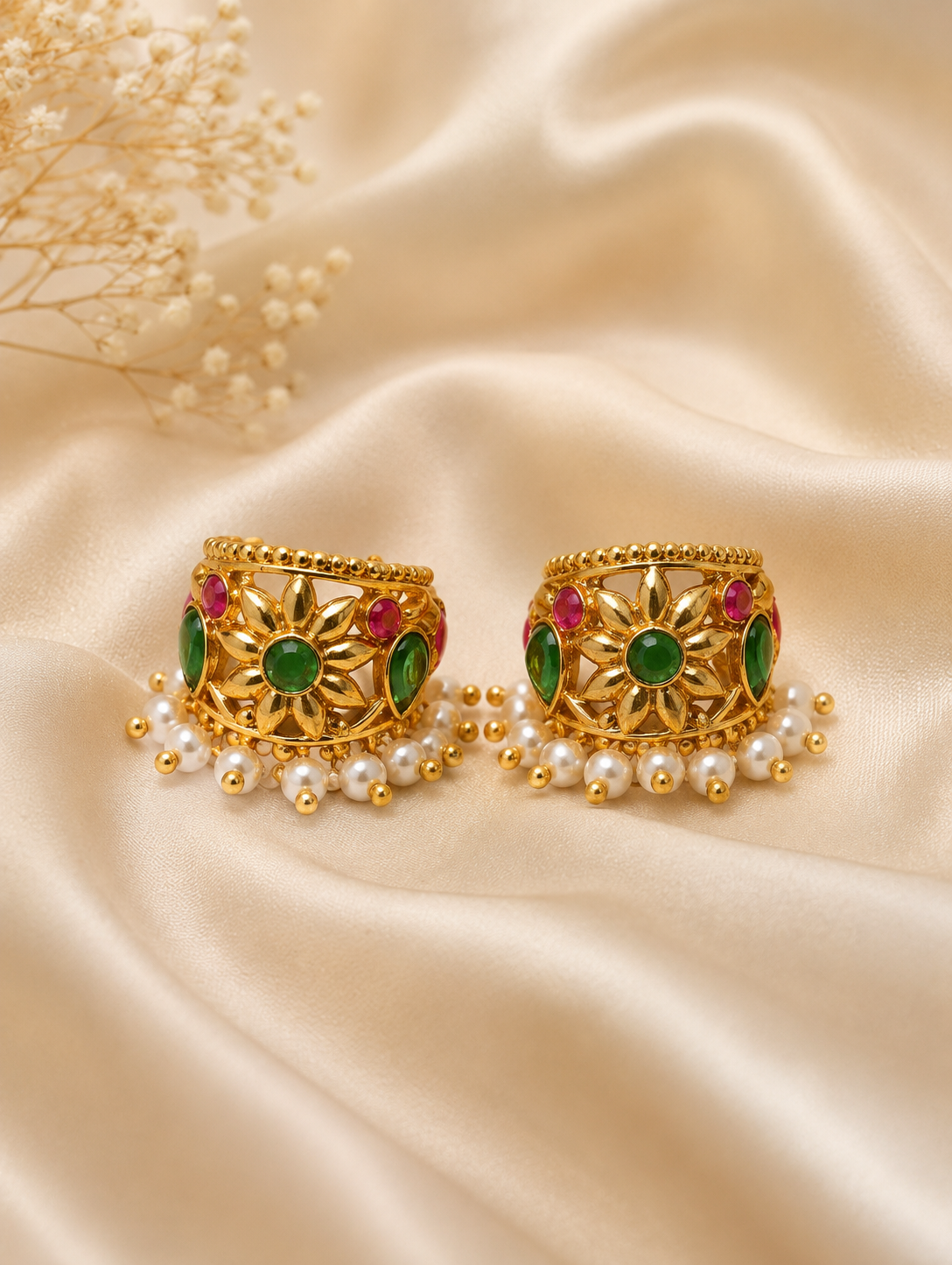 A pair of traditional gold-plated Bugudi cartilage earrings featuring a sculpted gold floral motif with a center emerald-green stone, accent ruby-red and green gems, and a fringe of pearl drops against champagne-colored silk fabric.