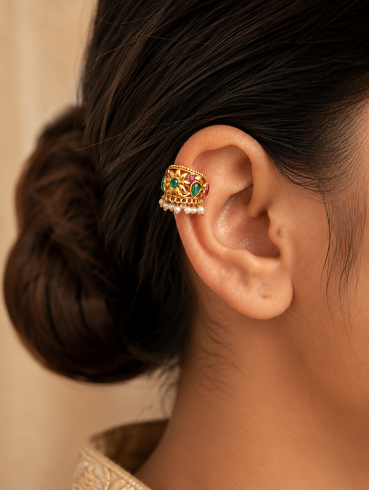 A pair of traditional gold-plated Bugudi cartilage earrings featuring a sculpted gold floral motif with a center emerald-green stone, accent ruby-red and green gems, and a fringe of pearl drops against champagne-colored silk fabric.