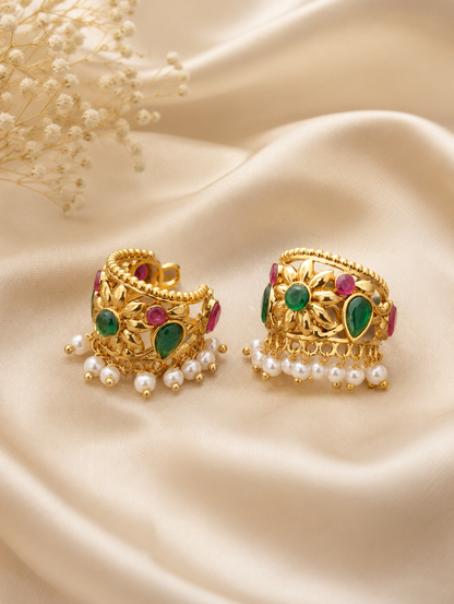 A pair of traditional gold-plated Bugudi cartilage earrings featuring a sculpted gold floral motif with a center emerald-green stone, accent ruby-red and green gems, and a fringe of pearl drops against champagne-colored silk fabric.