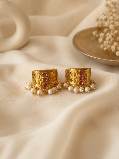 A pair of traditional gold-plated Indian Bugudi cartilage earrings featuring a geometric gold cutwork band adorned with a vertical row of three round ruby-red kemp stones and a bottom trim of white pearl drops, set against a draped off-white linen background.