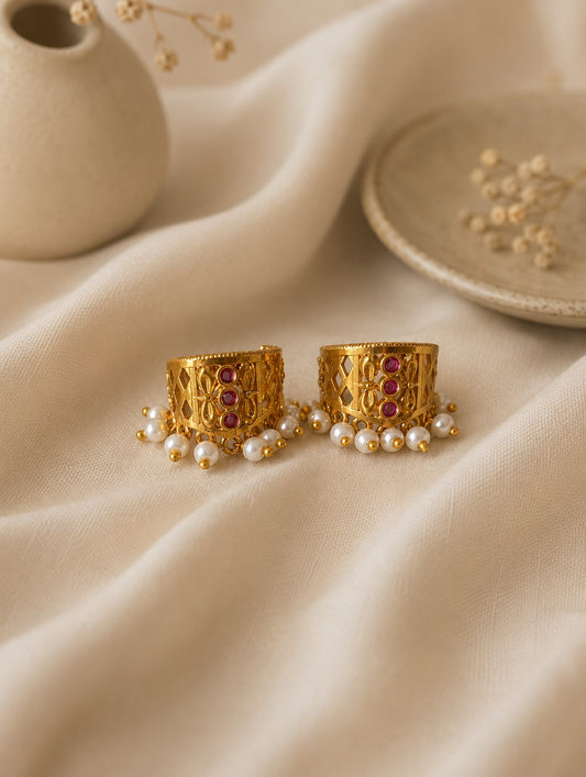 A pair of traditional gold-plated Indian Bugudi cartilage earrings featuring a geometric gold cutwork band adorned with a vertical row of three round ruby-red kemp stones and a bottom trim of white pearl drops, set against a draped off-white linen background.