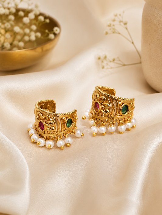 A pair of traditional Indian gold-plated Bugudi cartilage earrings featuring a central floral motif with an embedded round ruby-red kemp stone, accented by delicate cutwork and a bottom fringe of white pearls, resting on a draped cream satin fabric.