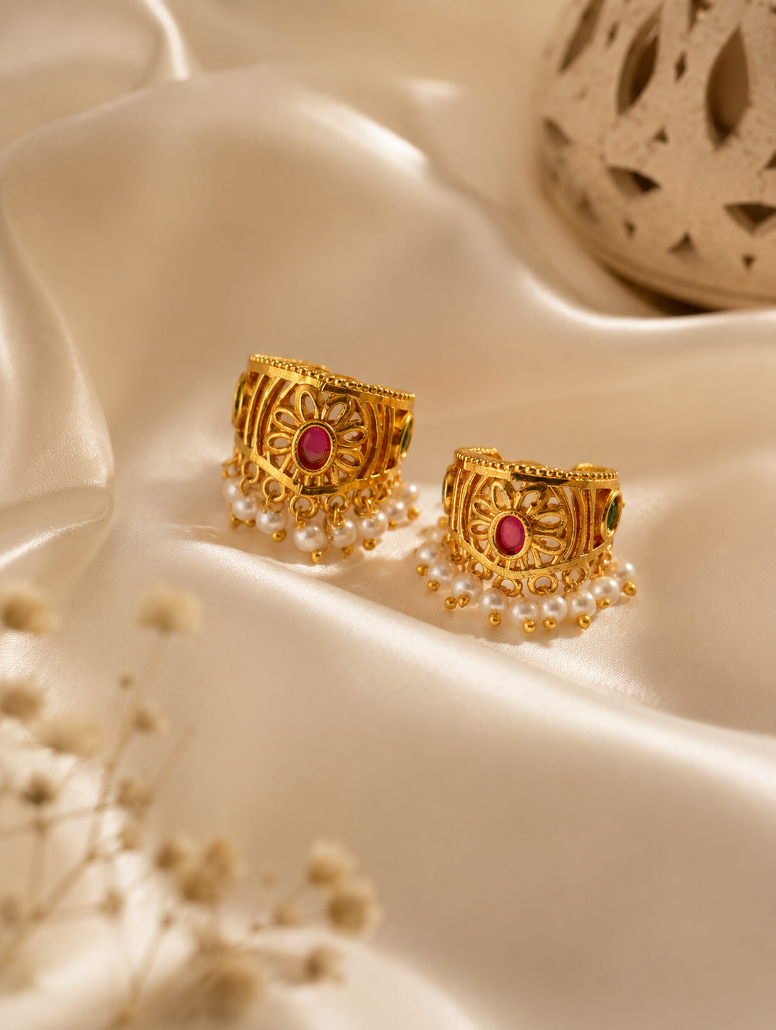 A pair of traditional Indian gold-plated Bugudi cartilage earrings featuring a central floral motif with an embedded round ruby-red kemp stone, accented by delicate cutwork and a bottom fringe of white pearls, resting on a draped cream satin fabric.