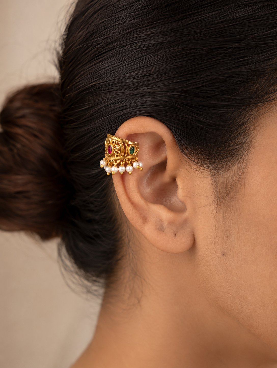 A pair of traditional Indian gold-plated Bugudi cartilage earrings featuring a central floral motif with an embedded round ruby-red kemp stone, accented by delicate cutwork and a bottom fringe of white pearls, resting on a draped cream satin fabric.