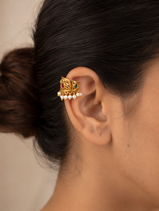 A pair of traditional Indian gold-plated Bugudi cartilage earrings featuring a central floral motif with an embedded round ruby-red kemp stone, accented by delicate cutwork and a bottom fringe of white pearls, resting on a draped cream satin fabric.