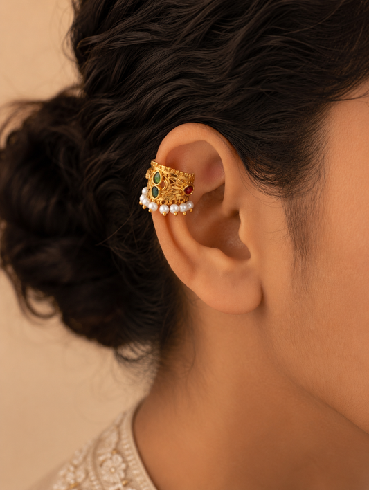 A close-up model view of a woman with dark hair styled in a bun, wearing a traditional Indian gold-plated Bugudi earring on her upper ear cartilage. The earring features green and red kemp stones, intricate gold detailing, and a delicate fringe of white pearls along the edge.