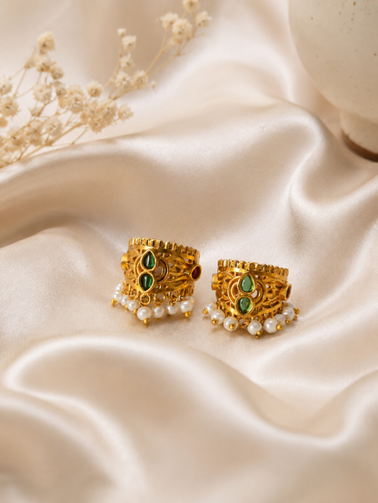 A close-up model view of a woman with dark hair styled in a bun, wearing a traditional Indian gold-plated Bugudi earring on her upper ear cartilage. The earring features green and red kemp stones, intricate gold detailing, and a delicate fringe of white pearls along the edge.
