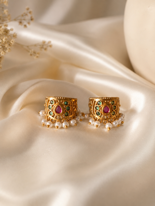 A close-up side profile view of a model with her dark hair styled in a neat bun, wearing a traditional Indian gold-plated floral Bugudi earring on her upper ear cartilage. The earring features vibrant kemp stone accents and a delicate trim of white pearls along the base.