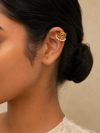 A close-up side profile view of a model with her dark hair styled in a neat bun, wearing a traditional Indian gold-plated floral Bugudi earring on her upper ear cartilage. The earring features vibrant kemp stone accents and a delicate trim of white pearls along the base.