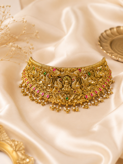 A broad antique gold temple jewelry choker necklace placed on a soft cream satin background, featuring an intricate relief carving of Goddess Lakshmi in the center surrounded by majestic peacock motifs, detailed with pink and green kemp stones, and finished with dangling small golden beads and pearls.