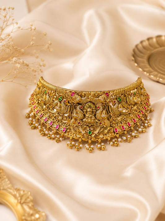 A broad antique gold temple jewelry choker necklace placed on a soft cream satin background, featuring an intricate relief carving of Goddess Lakshmi in the center surrounded by majestic peacock motifs, detailed with pink and green kemp stones, and finished with dangling small golden beads and pearls.