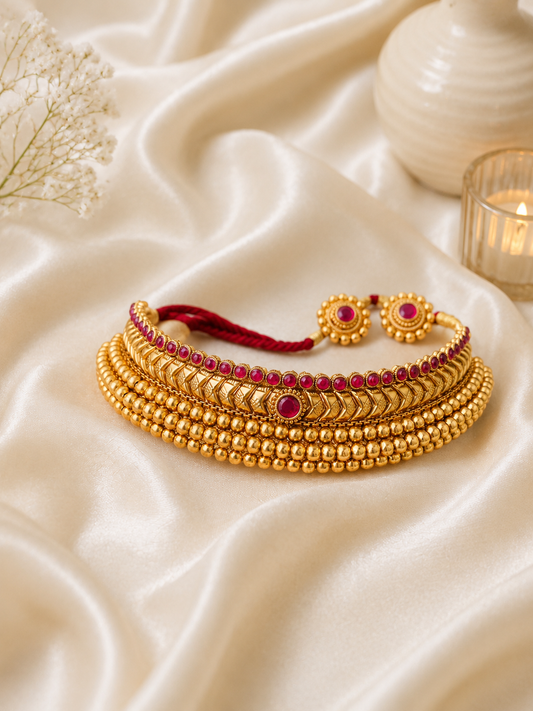 A traditional antique gold-plated Vajritika choker necklace arranged on a cream satin background, featuring multiple rows of polished golden beads, an intricate chevron patterned center band, and a top border lined with round pinkish-ruby stones, complete with an adjustable red thread cord.