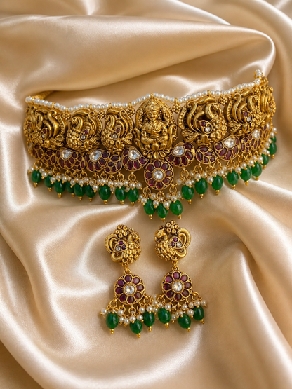 A grand antique gold temple jewelry choker and matching drop earrings displayed on a cream satin background. The choker features a center carving of a seated Goddess Lakshmi flanked by detailed peacocks, a row of tiny white pearls along the top, and a vibrant fringe of emerald green beads and small pearls at the bottom.