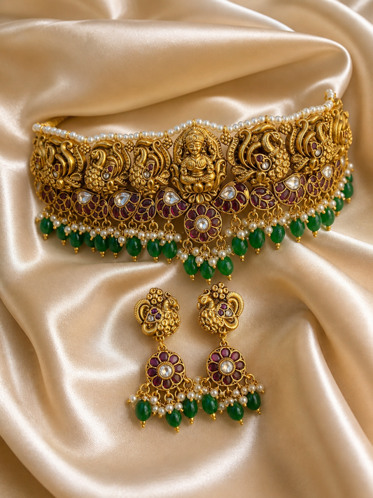 A grand antique gold temple jewelry choker and matching drop earrings displayed on a cream satin background. The choker features a center carving of a seated Goddess Lakshmi flanked by detailed peacocks, a row of tiny white pearls along the top, and a vibrant fringe of emerald green beads and small pearls at the bottom.