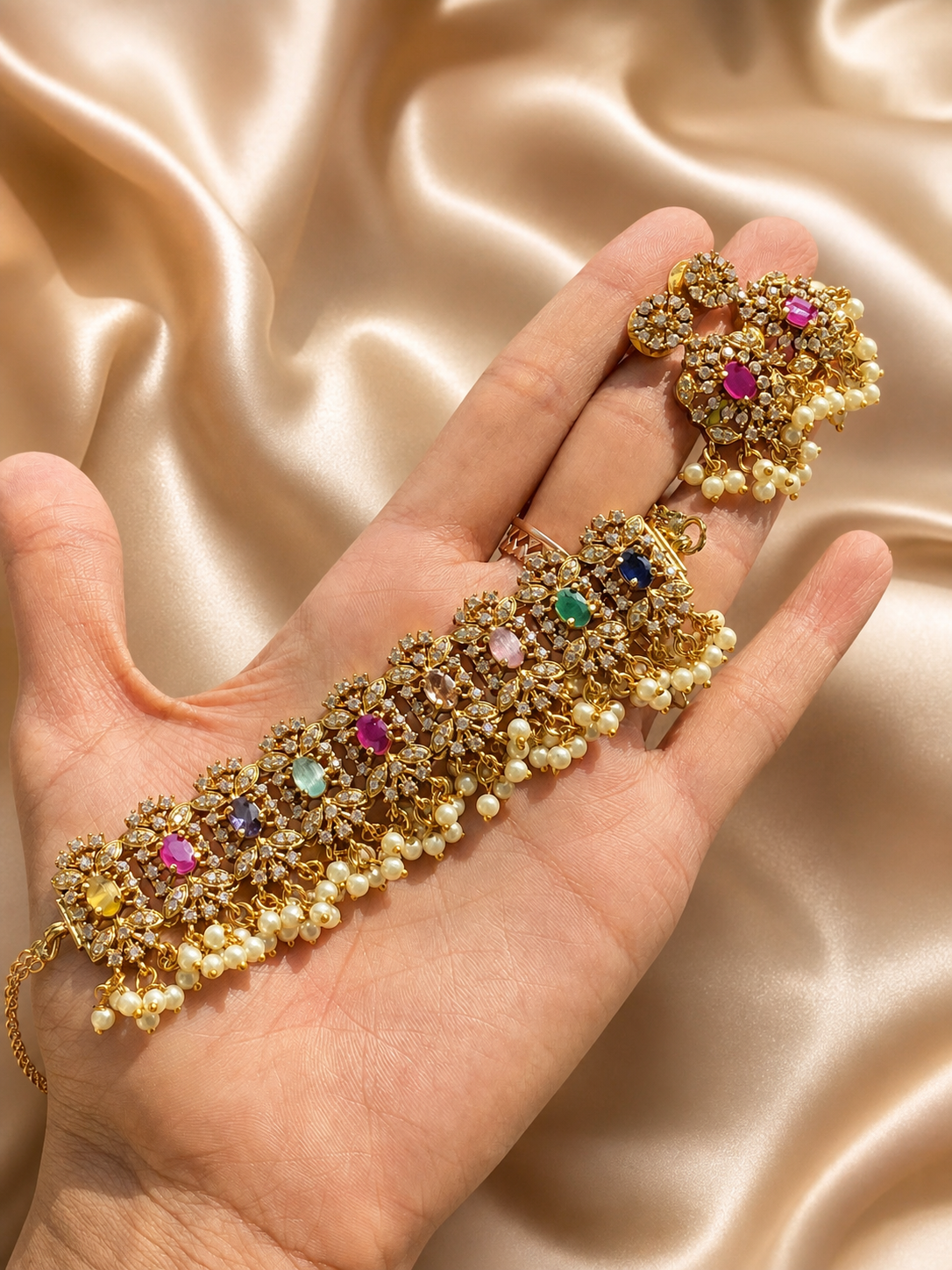 A hand holding a wide, multi-gemstone antique gold-plated choker necklace and matching statement earrings, featuring a lattice pattern studded with clear white stones, mixed colorful oval crystals, and a fringe of small hanging pearls on a champagne satin background.