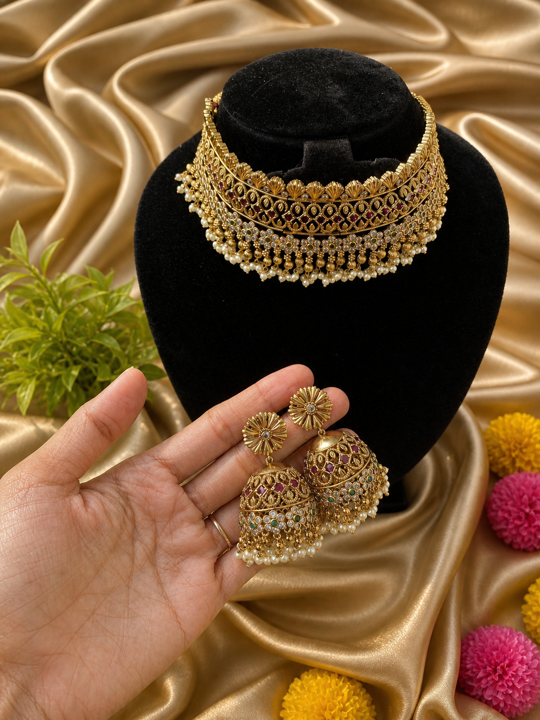 A broad, majestic antique gold-plated bridal choker necklace and matching jhumka earrings displayed on a soft cream satin background. The choker features a highly detailed traditional layout heavily studded with ruby red kemp stones, accented with a central green stone and a lush fringe of hanging clusters of small white pearls.