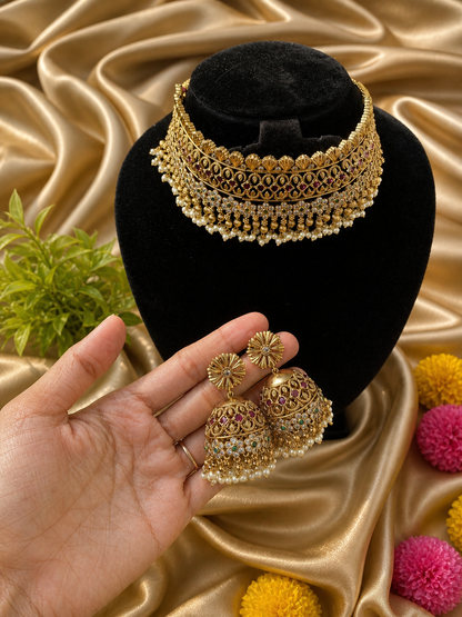 A broad, majestic antique gold-plated bridal choker necklace and matching jhumka earrings displayed on a soft cream satin background. The choker features a highly detailed traditional layout heavily studded with ruby red kemp stones, accented with a central green stone and a lush fringe of hanging clusters of small white pearls.