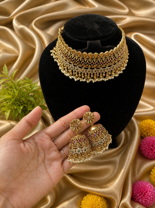 A broad, majestic antique gold-plated bridal choker necklace and matching jhumka earrings displayed on a soft cream satin background. The choker features a highly detailed traditional layout heavily studded with ruby red kemp stones, accented with a central green stone and a lush fringe of hanging clusters of small white pearls.