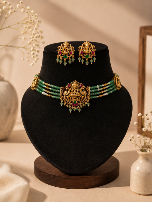 A beautiful temple jewelry set displayed on a black velvet bust, featuring a 4-strand choker made of light green beads and tiny pearls, centered around an antique gold-plated Goddess Lakshmi pendant with ruby red and emerald green accents, paired with matching statement stud earrings.