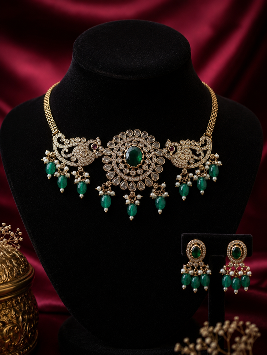 Haini traditional jewelry set featuring an antique gold-finish choker necklace with a central round emerald medallion flanked by stone-paved peacock motifs and hanging green teardrop beads, displayed on a black bust with matching drop earrings.