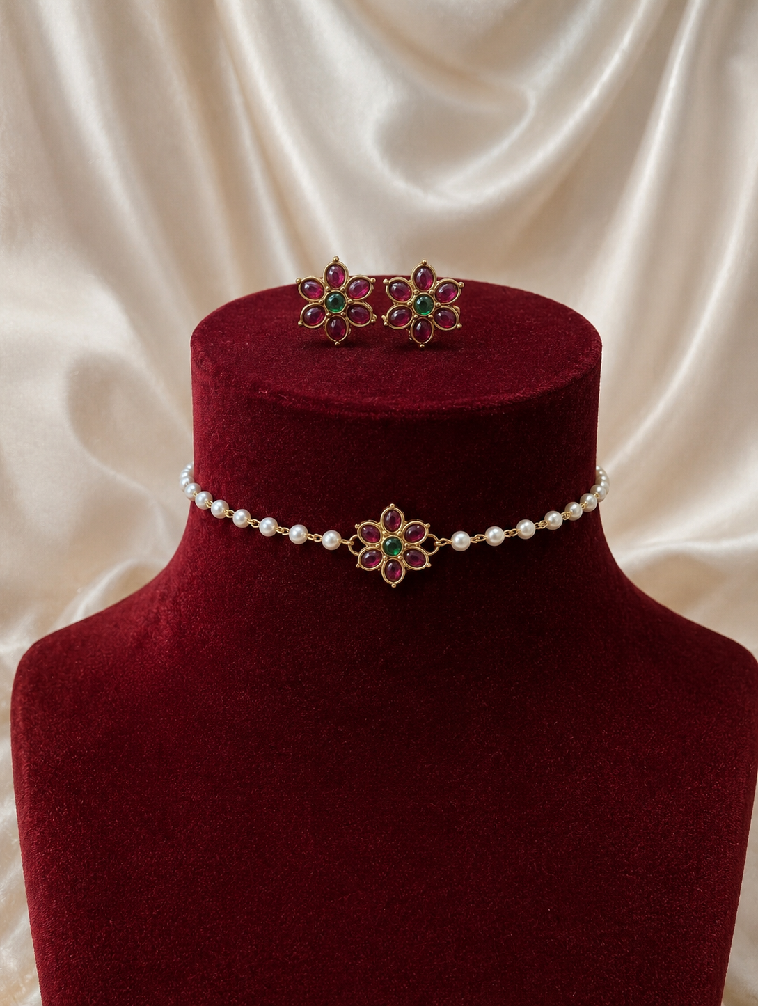 Traditional Malini jewelry set featuring a delicate white pearl choker necklace and matching stud earrings, both adorned with gold flower motifs, red rubies, and a green emerald center, displayed on a maroon velvet bust.