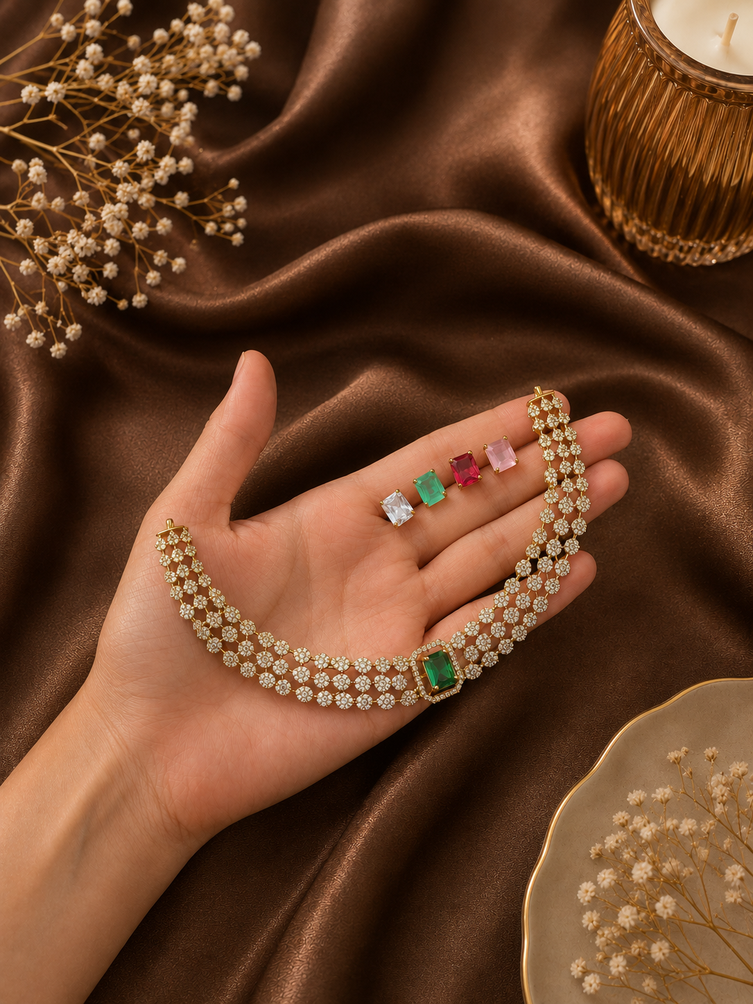A hand holding a three-row gold-plated floral CZ choker necklace featuring an interchangeable emerald green center stone, shown alongside alternative clear white, ruby red, and light pink rectangular replacement stones on a brown satin background.